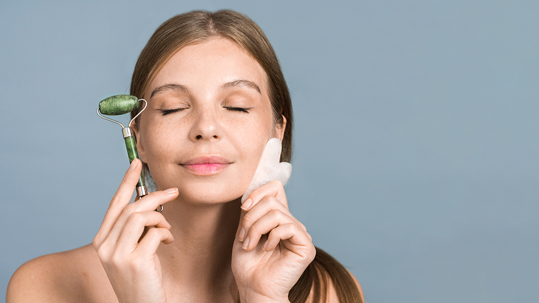 Young caucasian woman posing with roller and Gua Sha for facial massage